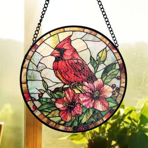 Puede incluir: Arte de ventana de vidriera redonda con un pájaro cardenal rojo posado en una rama con flores rojas y hojas verdes. La obra de arte está enmarcada en negro y cuelga de una cadena negra. El fondo es una mezcla de verde claro y amarillo.