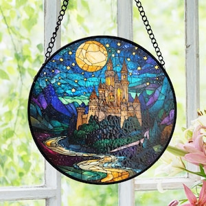 May include: Circular stained glass window art depicting a castle scene under a starry night sky and a full moon. The artwork has a black metal frame and chain. The castle is in gold and brown tones, with a river winding through the landscape.