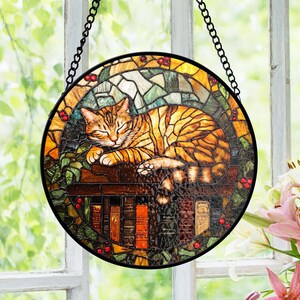 Op de afbeelding: Ronde glas-in-lood raamkunst met een slapende oranje tabbykat op een boekenplank. Het kunstwerk heeft een zwart metalen frame en ketting om op te hangen. Het ontwerp bevat groene bladeren, rode bessen en een kleurrijke achtergrond.