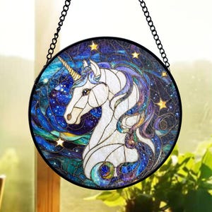 Può includere: Pannello rotondo in vetro colorato raffigurante un unicorno bianco con corno dorato e criniera viola. Lo sfondo è una galassia vorticosa di blu, verdi e viola, con stelle gialle. Una catena nera è attaccata per appenderlo.