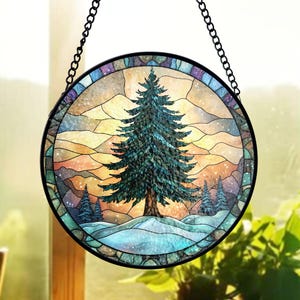 May include: Round stained glass window art featuring a winter scene with a large evergreen tree, snow, and mountains. The artwork is framed in black and hangs from a chain. The colors include blues, greens, yellows, and oranges.