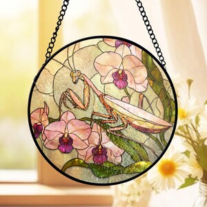 May include: Round stained glass window art featuring a praying mantis and orchids. The artwork has a black metal frame and chain for hanging. The mantis is tan and the orchids are pink and purple.