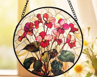 May include: A round stained glass window featuring a floral design with red and pink flowers, green leaves, and a cream-colored background. The window is framed in black and hangs from a black chain.