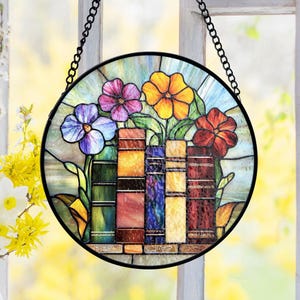 May include: A round stained glass window hanging with a black chain. The window features colorful flowers in shades of purple, orange, and red, arranged above a row of books. The books are in various colors, adding to the vibrant design.
