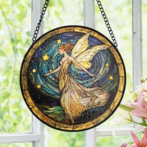 May include: Round stained glass window art featuring a fairy with outstretched arms and large yellow wings. The fairy is wearing a flowing gold dress, set against a swirling blue and gold starry background. The artwork is framed in black and hangs from a chain.