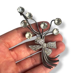 Op de afbeelding: Een zilverkleurige broche met een strik en een cluster van heldere strass stenen.