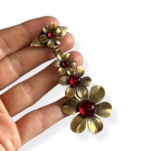 May include: A vintage floral brooch featuring four brass-toned flowers with red gemstone centres. The brooch has a leaf-shaped top and a chain of flowers that cascade down. The brooch is held in a hand against a white background.