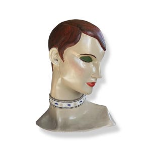 Puede incluir: Un busto de maniquí vintage con cabello rojo, sombra de ojos verde y lápiz labial rojo. El maniquí lleva un collarín de cuentas blanco con detalles azules y beige. La piel del maniquí tiene una tez pálida y pintada.