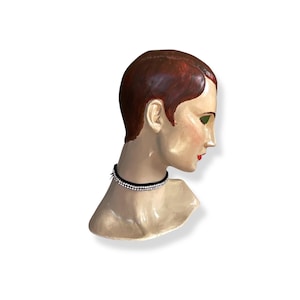 May include: A vintage mannequin head with red hair, green eye shadow, and red lipstick. The mannequin wears a black choker necklace with two rows of small, sparkling rhinestones. The head is painted with a vintage aesthetic.
