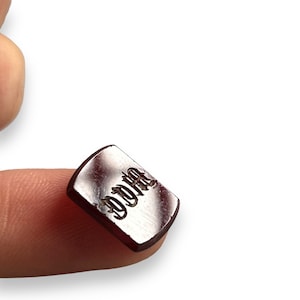 Può includere: Un piccolo ciondolo rettangolare in metallo color argento con la parola "DYNA" incisa in un font gotico. Il ciondolo è tenuto da un dito.