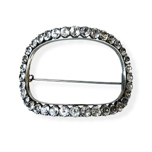 May include: An oval-shaped brooch with a silver-colored frame. The frame is lined with numerous clear, round gemstones. A pin extends from the back of the brooch for secure fastening. The brooch is set against a white background.