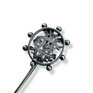 May include: A silver-toned metal hair pin with a circular decorative element. The circular design features an intricate pattern of interwoven shapes and is surrounded by small, spherical accents. The pin's stem is long and slender.