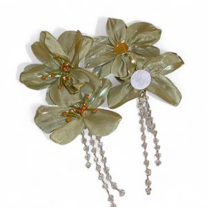 Op de afbeelding: Olijfgroene bloemen haaraccessoires met kralenaccenten en bungelende zilveren kettingen. De stoffen bloemen hebben een glinsterende, iriserende kwaliteit. Deze decoratieve haarstukken zijn ideaal voor speciale gelegenheden.