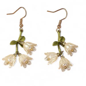 Può includere: Un paio di orecchini floreali con fiori bianchi a forma di campana e dettagli dorati. Gli orecchini hanno ganci color oro e steli verdi con foglie. Gli orecchini sono progettati per pendere e sono su sfondo bianco.