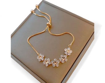May include: A gold-toned bracelet featuring a series of five flower-shaped charms. Each charm has a clear, sparkling center and is set in a gold-colored metal. The bracelet has an adjustable chain.