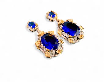 May include: A pair of gold earrings with blue gemstone accents. The earrings feature a large oval-shaped blue gemstone surrounded by a gold filigree design.