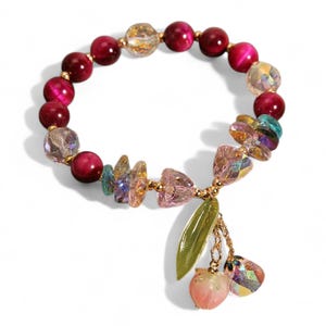 May include: A bracelet with deep red, round beads and iridescent and clear beads. It has a charm with a green leaf, a peach, and a crystal pendant. The bracelet is a piece of jewellery.