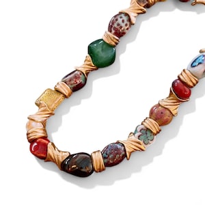 Op de afbeelding: Een ketting met een verscheidenheid aan kleurrijke, onregelmatig gevormde kralen. De kralen zijn in tinten rood, groen, bruin en wit en zijn verbonden door goudkleurige metalen accenten. De ketting wordt gepresenteerd tegen een witte achtergrond.