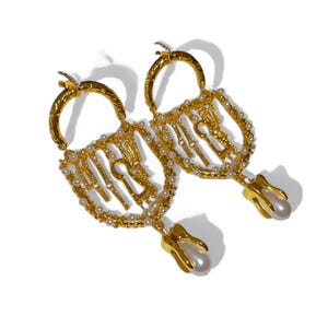 Könnte beinhalten: Ein Paar goldfarbene Ohrringe mit Schlüssel- und Schloss-Design. Die Ohrringe sind mit kleinen, runden, weißen Perlen verziert und haben eine hängende Perle am unteren Ende. Auf weißem Hintergrund.