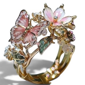 Op de afbeelding: Een gouden ring met een gedetailleerde vlinder met roze emaille vleugels en heldere steentjes, vergezeld door een roze en witte emaille bloem en groene bladaccenten. De ringband heeft een getextureerde gouden afwerking met kleine steentjes.
