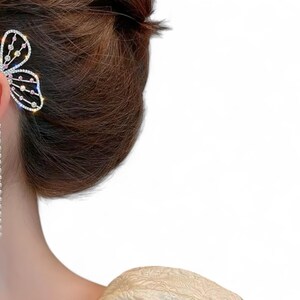 Peut inclure: Un ear cuff avec un motif de papillon orné de strass transparents. Le papillon s'enroule autour de l'oreille, avec des chaînes de strass pendantes. Une perle est attachée à l'oreille. Le bijou est argenté, sur des cheveux bruns.