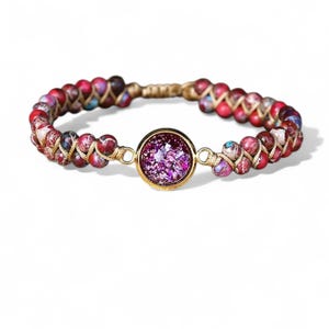 May include: A braided bracelet featuring red, pink, and purple beads with gold accents. The bracelet has a gold-colored center piece with a sparkling purple design. Jewelry.