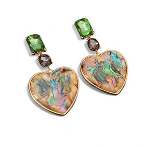 Puede incluir: Un par de pendientes en forma de corazón con borde dorado. Cada pendiente presenta una gema rectangular verde, una gema gris oscuro y un colgante iridiscente en forma de corazón. Los pendientes están sobre un fondo blanco.