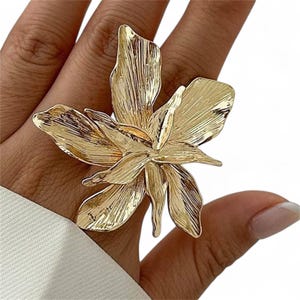 Può includere: Un anello a forma di fiore dorato con dettagliate texture dei petali. L'anello è un pezzo forte, caratterizzato da un ampio design floreale aperto. I petali hanno una finitura metallica strutturata, creando un look audace ed elegante. L'anello è progettato per essere indossato su un dito.
