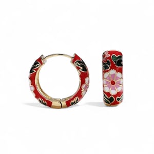 Op de afbeelding: Goudkleurige oorringen met een rood emaille bloemenpatroon. De oorbellen hebben een herhalend patroon van witte en roze bloemen met groene bladeren, tegen een rode achtergrond.