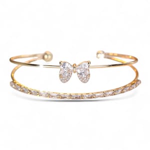 Puede incluir: Brazalete dorado con un centro en forma de lazo adornado con cristales transparentes. La banda inferior está adornada con cristales rectangulares. Una joya delicada.