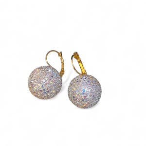 Può includere: Un paio di orecchini a leva con un design rotondo e scintillante. Gli orecchini hanno una montatura in metallo color oro e sono ricoperti di piccoli cristalli iridescenti. Gli orecchini sono esposti su sfondo bianco.