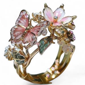 Könnte beinhalten: Ein goldfarbener Ring mit einem Schmetterling- und Blumendesign. Der Schmetterling hat rosa Flügel mit Kristallakzenten. Die Blüten sind rosa und weiß mit Kristallzentren. Grüne Blätter und Kristalle schmücken den Ring.