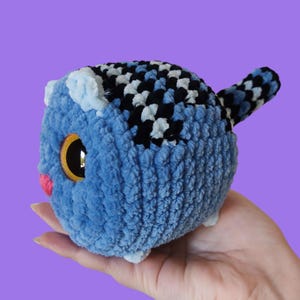 May include: A handmade, plush blue cat toy with a textured, fuzzy exterior. The cat has large golden eyes, a pink nose, and a black, white, and blue checkered pattern on its head and tail. The toy is round and has small white feet.