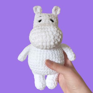 Puede incluir: Un juguete de hipopótamo blanco tejido a mano con pequeños ojos negros y orejas erguidas. El animal de peluche se sostiene sobre un fondo púrpura. El hipopótamo tiene un cuerpo redondeado, brazos pequeños y patas rechonchas.