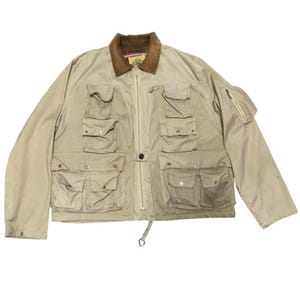 May include: A beige utility jacket featuring a brown corduroy collar, a full-length front zipper, and multiple front pockets with snap closures. The jacket includes a small zippered pocket on the left sleeve and a plaid lining visible at the neck.