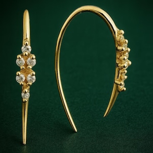 May include: Gold-toned earrings with a curved design. One earring features a vertical bar with five clear, round gemstones. The other earring has a curved hook shape with multiple gemstones. The earrings are set against a dark green background.