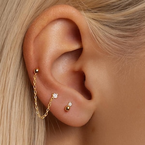 Puede incluir: Juego de aretes dorados. El juego incluye un arete de cadena con un pequeño diamante y un arete de botón con un pequeño diamante. También es visible un segundo arete de botón con una bola dorada.