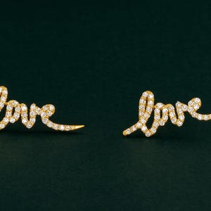 Może przedstawiać: Para złotych kolczyków z napisem "love" w formie kursywy. Kolczyki są inkrustowane małymi, błyszczącymi kamieniami szlachetnymi, na ciemnozielonym tle.
