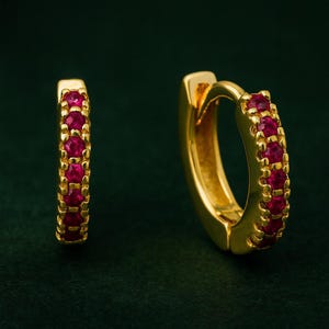 May include: Gold-plated hoop earrings featuring a line of small, vibrant red gemstones. The earrings have a polished finish and a classic design, suitable for everyday wear or special occasions. The gemstones are evenly spaced.