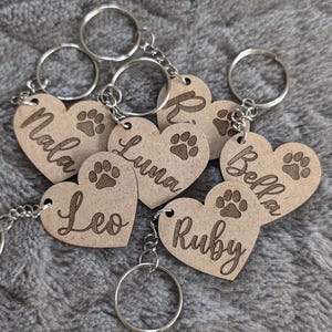 May include: Six wooden heart-shaped keyrings with engraved names and paw prints. The names are "Mala", "Leo", "Luna", "Bella", "Ruby", and "Kiki".