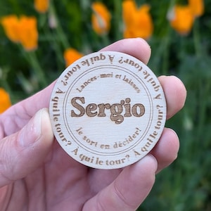 May include: A wooden token with the name "Sergio" engraved on it. The token has text in French that reads "A qui le tour?" which translates to "Whose turn is it?"