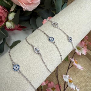 May include: Silver-toned bracelets with various charms, including an eye design with blue and clear stones. The bracelets are displayed on a white, cylindrical surface, with pink flowers in the background.