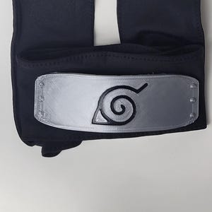 May include: Black headband with a silver metal plate featuring a spiral design. The headband is made of a dark fabric and has two long, rectangular extensions. The metal plate has a textured appearance.