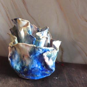 May include: A ceramic vase with a distinctive, ruffled design. The vase has a mix of blue, white, and tan colours, creating a marbled effect. The edges of the vase are folded and textured, enhancing its artistic appeal. The vase is a decorative item.