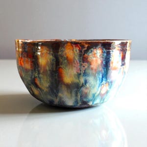 May include: A round ceramic bowl featuring a glossy, multi-coloured glaze with swirling patterns of deep blue, vibrant orange, yellow, and dark brown. The surface has a reflective finish with a smooth, curved rim and a rounded base structure.
