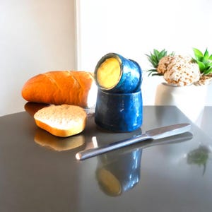 May include: A blue ceramic butter dish with a silver butter knife, a loaf of bread, and a slice of bread. The butter dish contains yellow butter. The items are on a reflective surface.