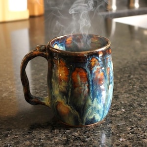 May include: A handcrafted ceramic mug with a distinctive glaze in shades of blue, orange, and green. Steam rises from the mug, suggesting a hot drink. The mug has a robust handle and a textured surface.