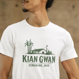 Puede incluir: Camiseta blanca con un gráfico verde de un edificio, palmeras y un barco, con el texto "KIAN GWAN SEMARANG, JAVA". La camiseta la lleva una persona.