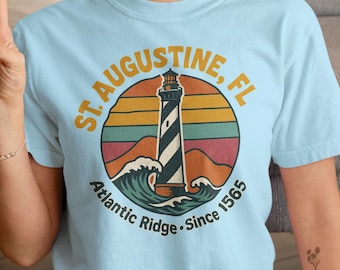 May include: Light blue t-shirt with a retro-style graphic of a lighthouse in front of a sunset. The graphic says "St. Augustine, FL Atlantic Ridge - Since 1565".