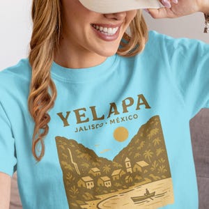 Puede incluir: Camiseta azul claro con una ilustración marrón de Yelapa, Jalisco, México. El diseño muestra un río, montañas, edificios y el texto "Yelapa Jalisco Mexico". La persona de la imagen lleva una gorra beige.